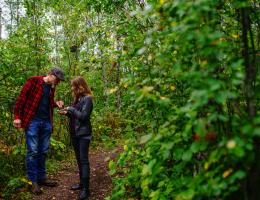 two visitors examine something in the woman's hand standing in the leafy green trees.