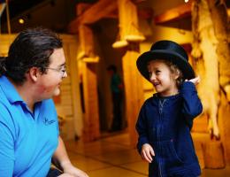 An historical interpreter smiles at a young visitor who is trying on the top hat.
